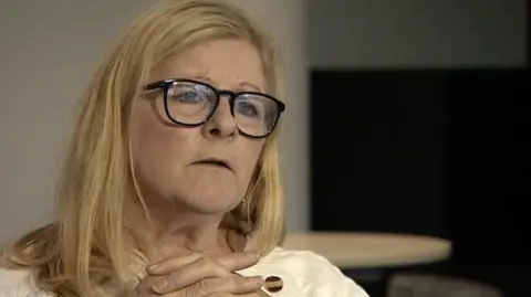 BBC A woman with blonde hair wearing glasses with her hand crossed at collarbone level. She has black-rimmed glasses and sits in a room with white and dark walls and a round, wooden table behind her.