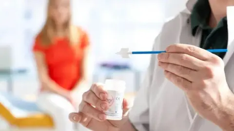 Science Photo Library A doctor in white scrubs holding a cervical smear test. A woman in white trousers and an orange top sits behind her, blurred out in the background.