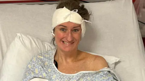 Susannah Morgan Susannah is propped up in a hospital bed with a bandage around her head. She is wearing a hospital gown and is attempting to smile but one side of her mouth is drooping down.