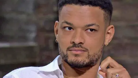 BBC Steven Bartlett pictured on the set of Dragons' Den. He has short hair with a fade on the side, and a short beard. He is wearing a white, open necked shirt, small hooped and stud earrings, a gold chain necklace and a gold ring studded with coloured gems. Behind him is an exposed brick wall. 