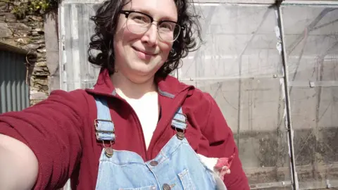 HANDOUT Alix Harvey, wearing glasses and a red fleece with denim dungarees, smiles in a selfie 