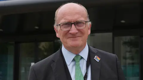 Nikki Fox/BBC Nick Hulme in a black suit jacket with a blue shirt and a green tie. He is wearing glasses and is looking into the camera and smiling.