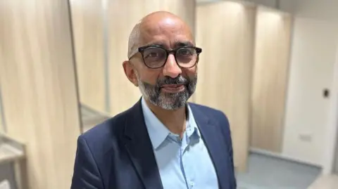 Dr Saket Priyadarshi smiles at the camera. He is bald with a black and grey beard. He wears a navy suit with a light blue short and black glasses.