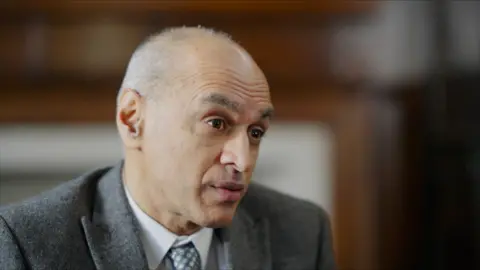 Prof Naveed Sattar, a man with closely cropped grey hair, wearing a grey flannel suit and a silk tie with a pattern of grey dots, speaking to someone on the right of the camera as he sits in a wood-panelled room.