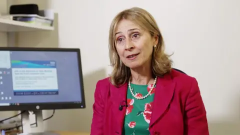 Professor Barbara McGowan, a woman with blonde hair, who is wearing a fuchsia jacket and a green dress with a pattern of pink flowers, sitting in a hospital consulting room with her computer screen beside her.