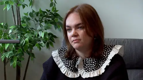 Emily Dove sits in a chair. She is wearing a navy blue top with a polka dot-patterned collar. She has shoulder-length brown hair and a pierced nose.