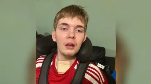 Hilary Rabbett George Rabbett, wearing a chest harness and red t-shirt, looks into the camera. There is a large plaster on his throat.