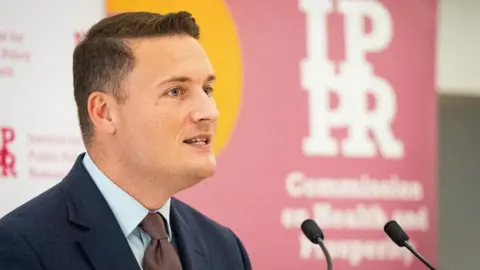 PA Media A side profile of Health Secretary Wes Streeting stood at a podium speaking into a microphone wearing a navy suit, blue shirt and brown tie. He is clean shaven with short, dark hair.