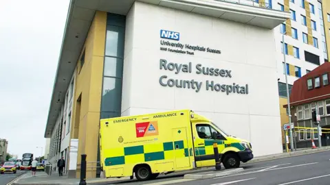 Mark Norman / BBC A yellow ambulance enters an access road on a slope, alongside a modern building with the words 