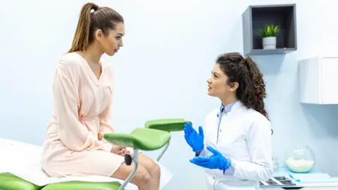 Getty Images Stock photo of female gynecologist wearing blue medical gloves talks to a female patient wearing a peach coloured dress who is sat on a medical bed that has stirrups attached to the end