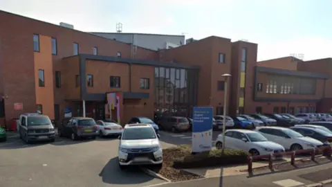 Google The outside of a hospital, with a car park in front of it. The hospital building is red brick with black frames around the centre structure. 