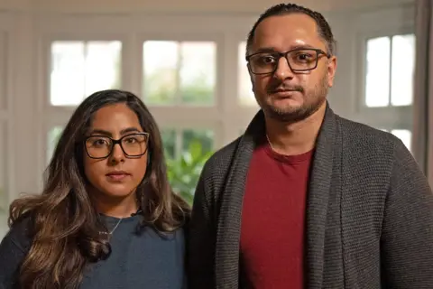 MARTIN MCQUADE / BBC Amarjit and Mandip pictured standing next to each other. Amarjit has long brunette hair and black-rimmed glasses. She is wearing a blue jumper and a silver necklace. Mandeep has dark hair which is tied back, black-rimmed glasses and a short beard. He is wearing a red t-shirt and grey woollen cardigan. They are pictured in front of a white-framed window with green plants outside. 