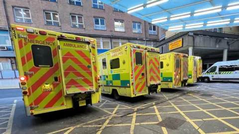 BBC The outside of an Emergency Department main entrance. 5 ambulances are parked outside.