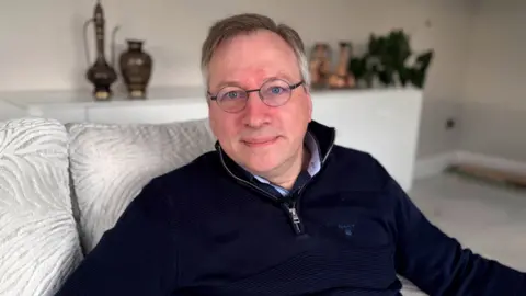 BBC A man with round glasses and short grey hair sits on a silver sofa, looking down the lens of the camera. He has his lips pursed and his eyes are a piercing blue colour. He is wearing a navy quarter zip jumper with a silver zip.