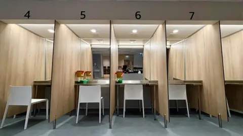 BBC Numbered booths with a white chair in each booth. Each one faces a mirrored wall. It is brightly lit.