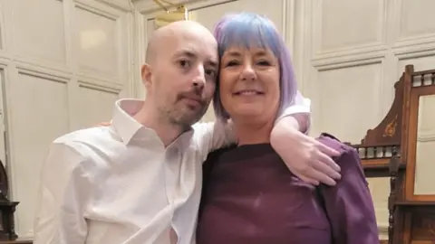 Roisin Wilshaw A man and a woman are looking at the camera - the man, who is bald with light facal hair and wearing a white shirt, is on the left. The woman, whose hair is dyed blue and violet, is on the right. She is wearing a burgundy top. In the background are white walls. 