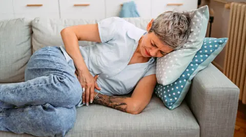 Getty Images A woman with short hair lies on a sofa, clutching her stomach. She has short grey hair and wears a blue shirt with blue jeans. 

The sofa is grey. 