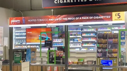 Tobacco kiosk at a Morrisons store showing adverts for two different brands of heated tobacco, Ploom and iQos, behind a perspex screen. Ploom slogan reads 