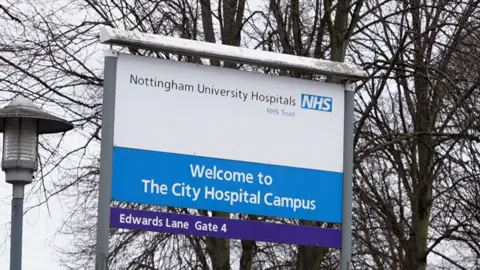 A blue and white sign outside Nottingham's City Hospital which says: Welcome to The City Hospital Campus, Nottingham University Hospitals NHS Trust.
There is a street light to the left of the sign and trees with no leaves on, behind the sign.