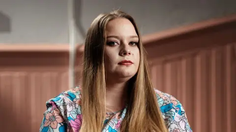 Abby sits and looks at the camera. She has long light brown hair and a floral dress.