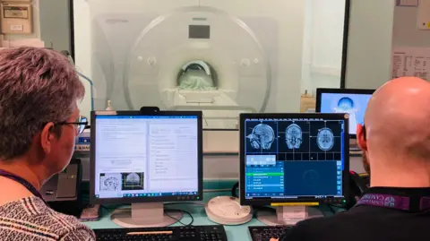 Two radiographers - one female and one male - are looking at MRI images of Samantha's brain on three computer screens. They are photographed from behind so the screens are visible. In the distance, behind a glass partition, is an MRI scanner.