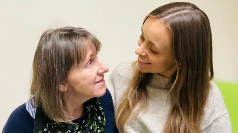 BBC Patient Samantha Dennison is sitting on a hospital bed and looking at her daughter Steph. Samantha has shoulder length brown hair and wears a dark blue cardigan over a floral top. Steph has long brown hair and wears a light grey,  high-neck jumper.