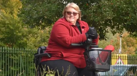 Sue Myerscough A woman in a red fleece coat is sitting on a mobility scooter.  She has shoulder length, light brown hair, and is wearing sunglasses.  She is on a path in the middle of a park and is surrounded by trees and greenery.