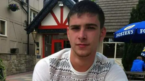 Jayne Donn A photo of James sitting in a beer garden outside a pub. He has short dark hair and is clean-shaven. He is looking directly at the camera, wearing a white jumper with a pattern on the front but not the sleeves.