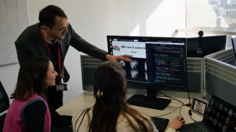 King's College London One male researcher stands, pointing at a computer screen, while two female researchers sit in front of the screen, with one holding a computer mouse.