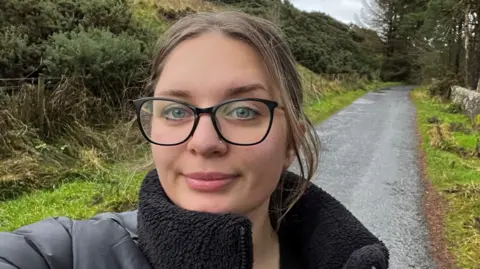 Weronika Somerville Weronika takes a selfie on a country lane, with a path leading off behind her and hedges on her right side, a row of trees on her left. It's a damp day and she is wearing a black puffer jacket with a fleece lining. She is also wearing stylish large-framed glasses, her light brown hair pulled back in a pony tail.