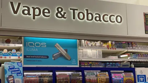 BBC Vapes and tobacco kiosk in a Sainsbury's store in London. Sign reads 'vape and tobacco'. Below is a blue sign advertising iQos Iluma, 