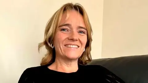 Sex Matters Sandie Peggie, a woman with shoulder-length blonde hair, smiles at the camera while sitting on a couch. She is wearing a black top.