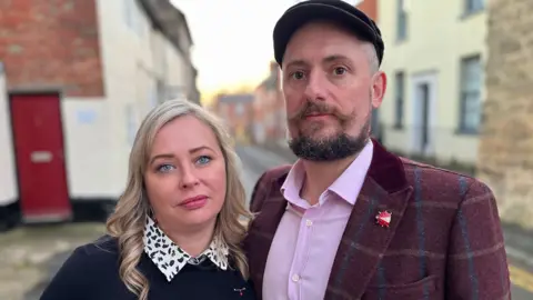 BBC Couple Carly and Carl are pictured standing together in the street. There are terraced houses in the background. Carly has blonde hair and is wearing a black and white shirt and a dark jumper. Carl is wearing a cap and has a beard and moustache. He is wearing a lilac shirt with a maroon checked jacket. He has a ladybird brooch on the lapel.