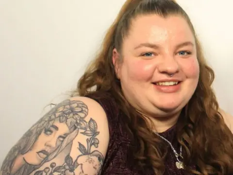 Jess Phillips A woman with wavy brown hair and tattoos on her arm smiles at the camera