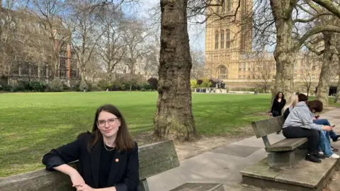 Transport for All chief executive Caroline Stickland sat on a park bench with Westminster in the background.