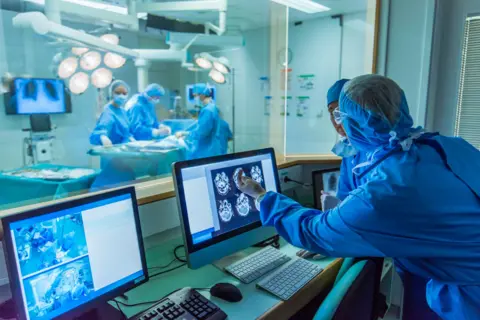 Getty Images Stock photo of people looking at brain images during an operation. In the background is an operating theatre with four staff and a patient on the table. This is blurred. In the foreground are four people dressed in blue surgical scrubs looking at computer screens with brain scans on them and one is pointing at one. 