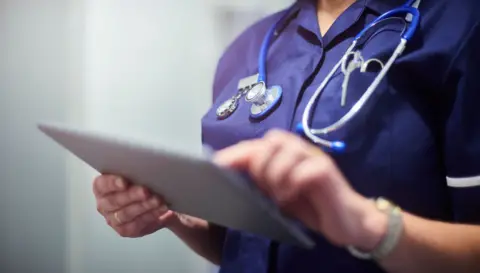 Getty Images Close-up stock picture of a nurse in navy blue uniform with stethoscope around her neck and watch clipped to her uniform. She is holding a clipboard and pointing at something on it. We cannot see her face. 