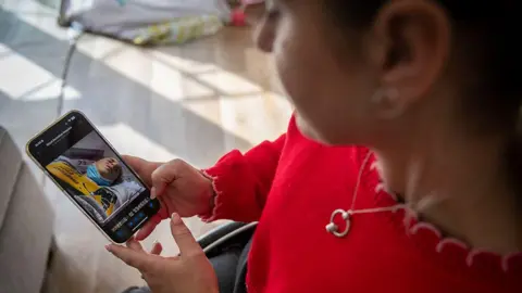 BBC / Emma Lynch A picture taken over Dani's shoulder shows her looking down at her phone, on which a video is playing showing her lay down in a hospital bed, wearing her ice hockey kit. She has a face mask pulled down over her chin and her face is twisted in pain.
