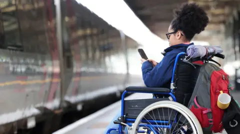 Getty Images Female wheelchair user waiting on a train platform, using her phone.