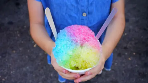 Getty Images A child wearing a blue dress holding a multi-coloured slushy ice drink out in front of them with both hands around it.