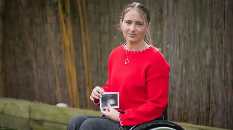 BBC / Emma Lynch Dani is in her wheelchair holding a scan photo of the child she had to abort on medical grounds. Dani is looking directly into the camera and is wearing a red jumper.
