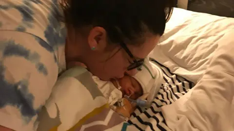 PA Media Sarah Robinson, who has dark brown hair pinned up and brown glasses, kisses her baby daughter Ida Lock on the cheek. The baby girl is wearing a white hat and is attached to tubes and wires.