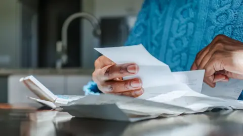 Getty Images Close up shot of a person looking at bills and receipts on a table next to a calculator in their kitchen. They are wearing a blue jumper and the kitchen sink is in the background.