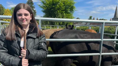 Julia Gregory/ BBC A young woman with shoulder length dark hair and and wearing a black jacket is standing in front of a pen with cows inside. None of them are looking at the camera. A church spire can be seen in the background.