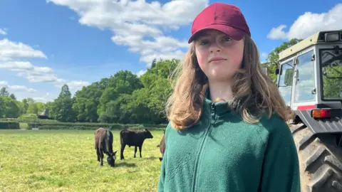 Julia Gregory/ BBC Dressed in a green top and red cap a teenager stands beside a tractor with large wheels. several cows are in the background and one is grazing the grass.