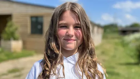Nancy, aged nine, smiling wearing a white school polo shirt with shoulder length, light brown hair. She is outside and while the background is blurred, she appears to be standing in front of stables.