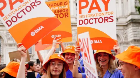 PA Media Junior doctors protest in August 2023, wearing blue scrubs and orange hats hold placards reading: