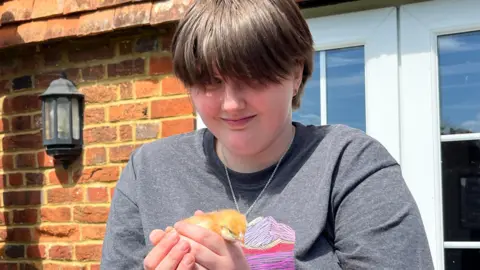 Julia Gregory/BBC A young woman carefully holds a week-old yellow chick in both her hands. She is smiling at the camera and has short dark hair and a black T-shirt.
