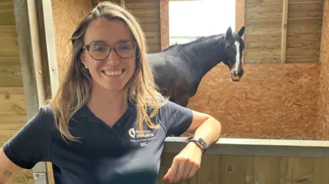 Mariachiara Apruzzese smiling wearing a navy blue Horses for Wellbeing polo shirt leaning on a stable door with her favourite horse, 