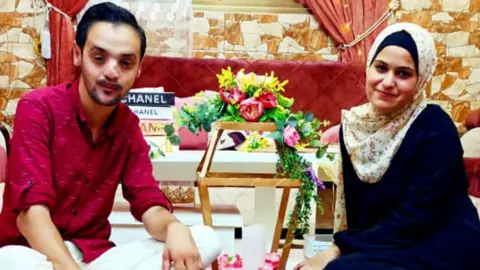 Noura's family archive Mohamed and Noura sitting down in their home with some flowers between them. He is wearing a red shirt and she is wearing a dark blue dress and light patterned headscarf.